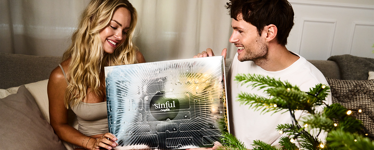 Ett leende par som håller i en Sinful-julkalender bredvid en pyntad julgran