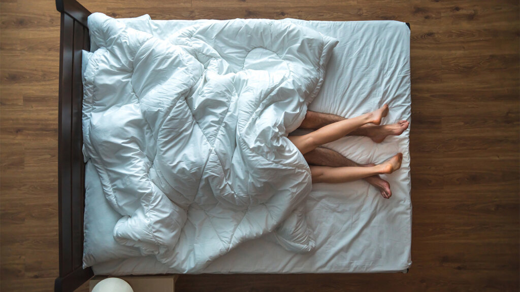 Two people lie in a bed with the duvet over their faces and most of their bodies