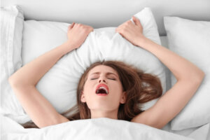 Kvinne som ligger i en seng under dynen med hendene over hodet som holder i puten
