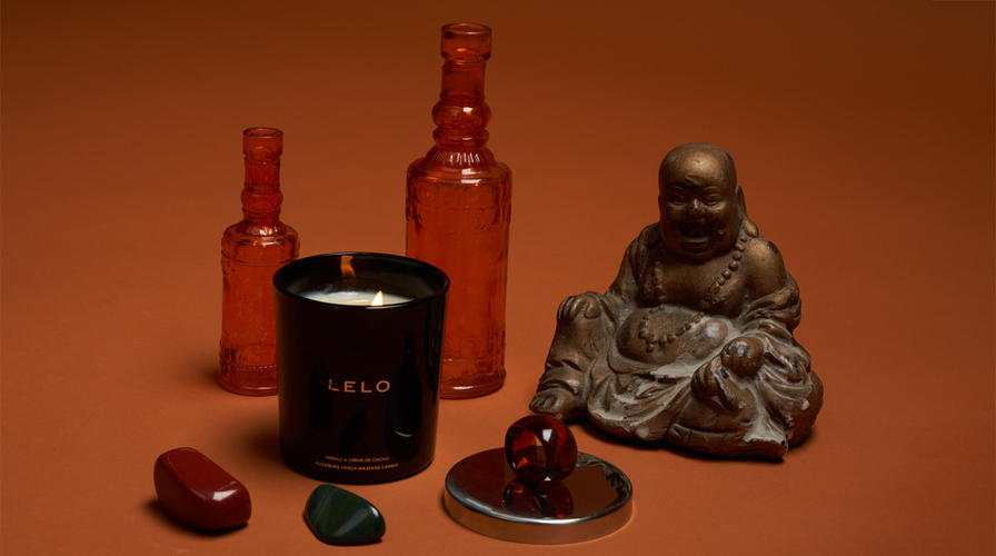 Une bougie de massage Lelo, des cristaux, des bouteilles décoratives en verre et un Bouddha en bronze sur un fond orange