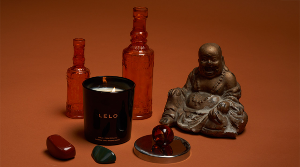 Et lille LELO massagelys, krystaller, glasflasker og en Buddha statue i bronze med en orange baggrund