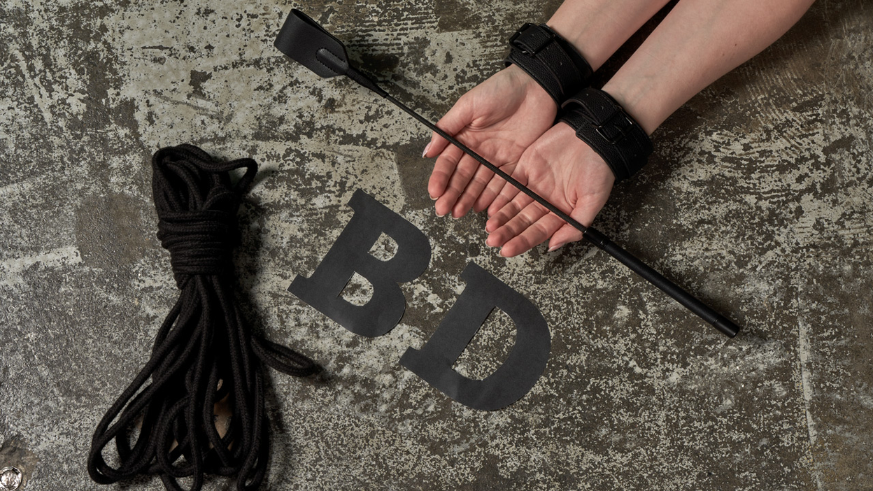 The letters BD, a bondage rope and two hands holding a whip