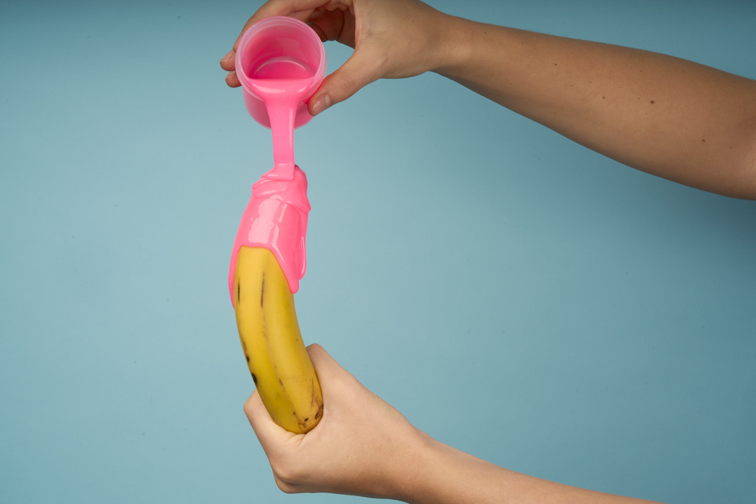 Nærbillede af to hænder der hælder lyserødt silikone ud over en banan