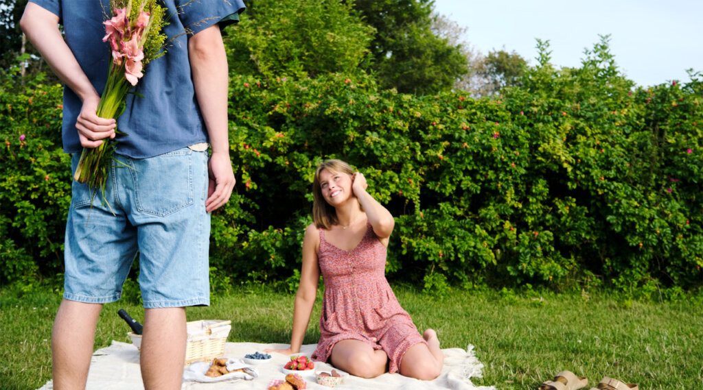 Henkilö pitää kukkapuskaa selkänsä takana piilossa piknik-viltillä istuvalta kumppaniltaan