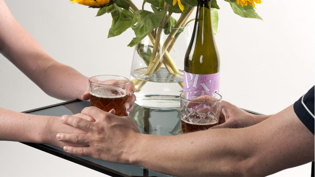 Två personer händerna över ett bord med två glas öl och en vas med solrosor