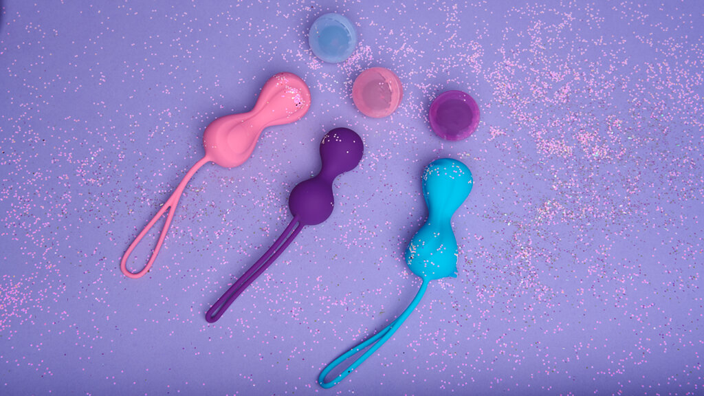 Three kegel balls in different colours on a light purple background with glitter