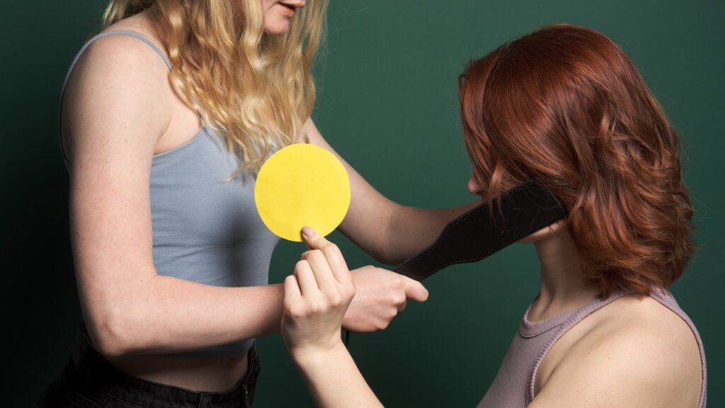 Vrouw laat een geel bord zien terwijl een andere vrouw een peddel tegen haar wang houdt 