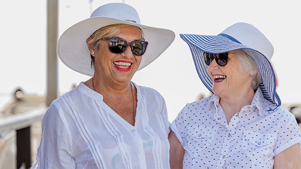 Δύο γυναίκες, χαμογελόντας η μια στην άλλη, φοράνε καπέλο και γυαλιά ηλίου