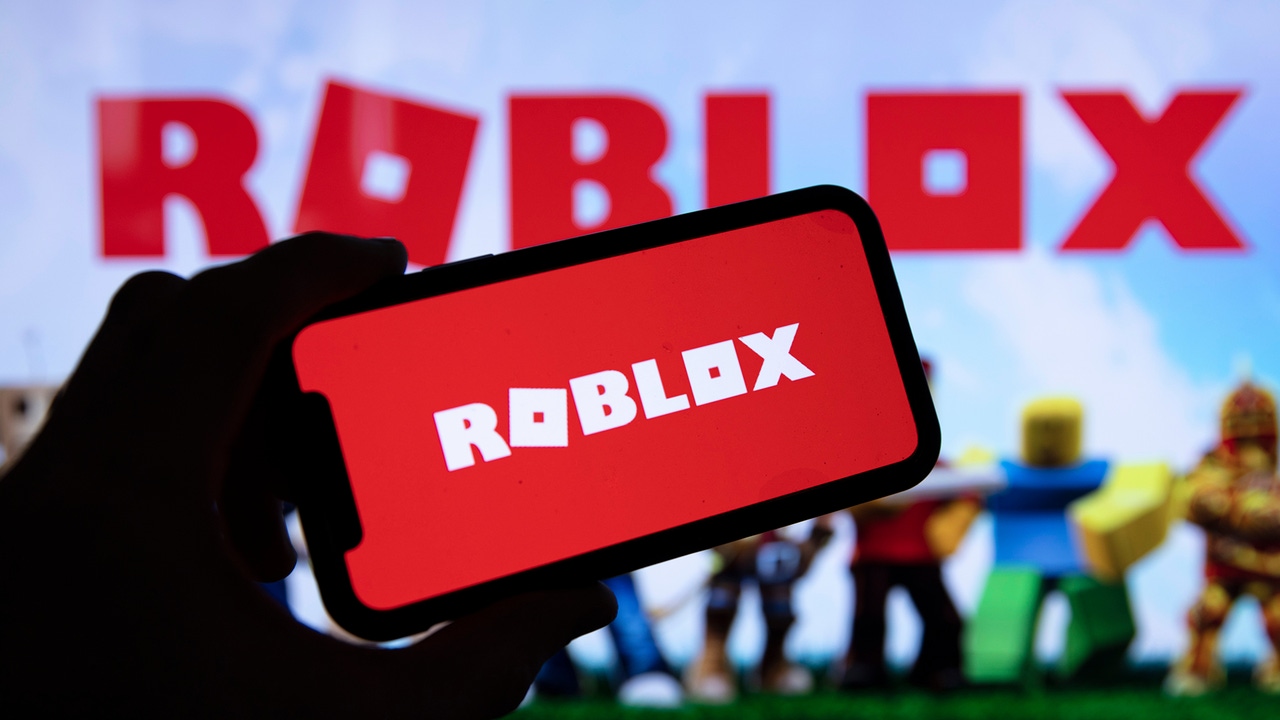 A silhouetted hand holds up a phone with the Roblox photo on it against key art for Roblox A silhouetted hand holds up a phone with the Roblox photo on it against key art for Roblox