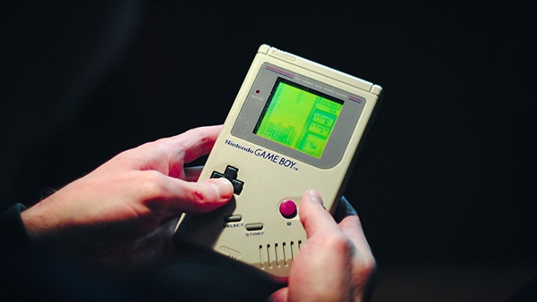 A pair of hands hold a classic Game Boy. A game of Tetris is onscreen.