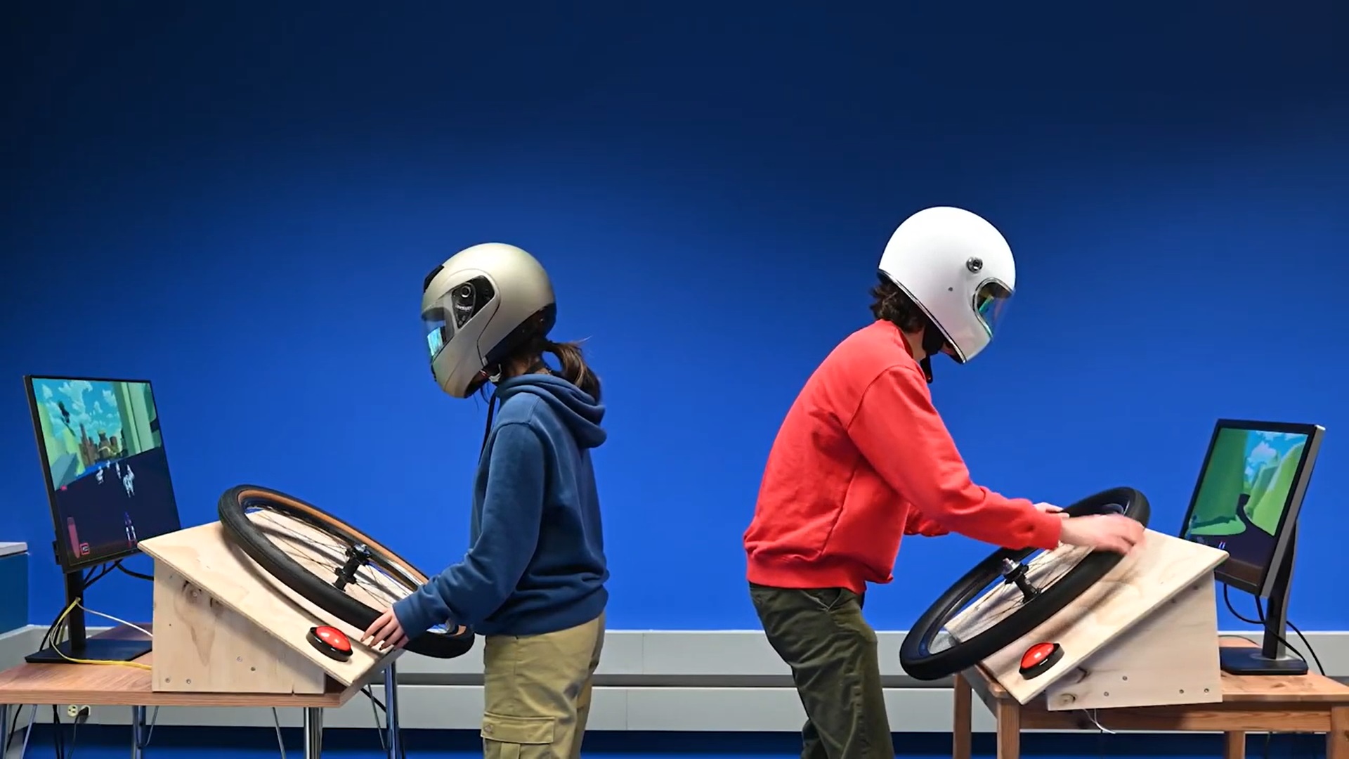 two people in racing helmets sitting back to back, each steering using a bicycle tire steering wheel