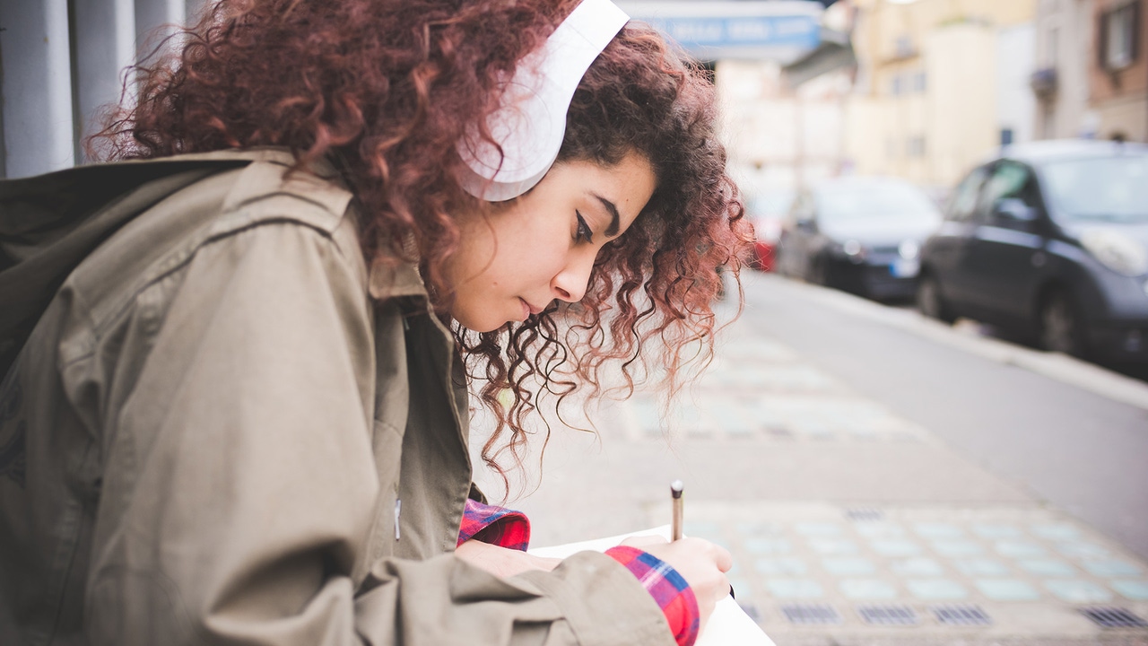 A woman with curly hair sketches while sitting on a curb. A woman with curly hair sketches while sitting on a curb.