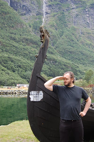 The developer saluting outside of a beached Norwegian ship, with the fjords and a body of water visible in the background.