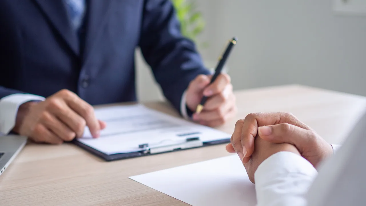 A man in a blue suit presents papers to a man in a white shirt clasping his hands. A man in a blue suit presents papers to a man in a white shirt clasping his hands.
