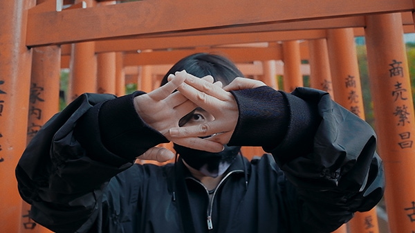 A still frame of Ikumi Nakamura. She makes a symbol with her hands while her face is concealed behind a black face mask.