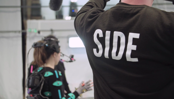A person in the foreground with the word SIDE printed on their back holds a boom mic toward a woman in a motion capture suit.