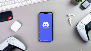 A phone with the Discord logo on it next to two PS5 controllers and a keyboard. A phone with the Discord logo on it next to two PS5 controllers and a keyboard.