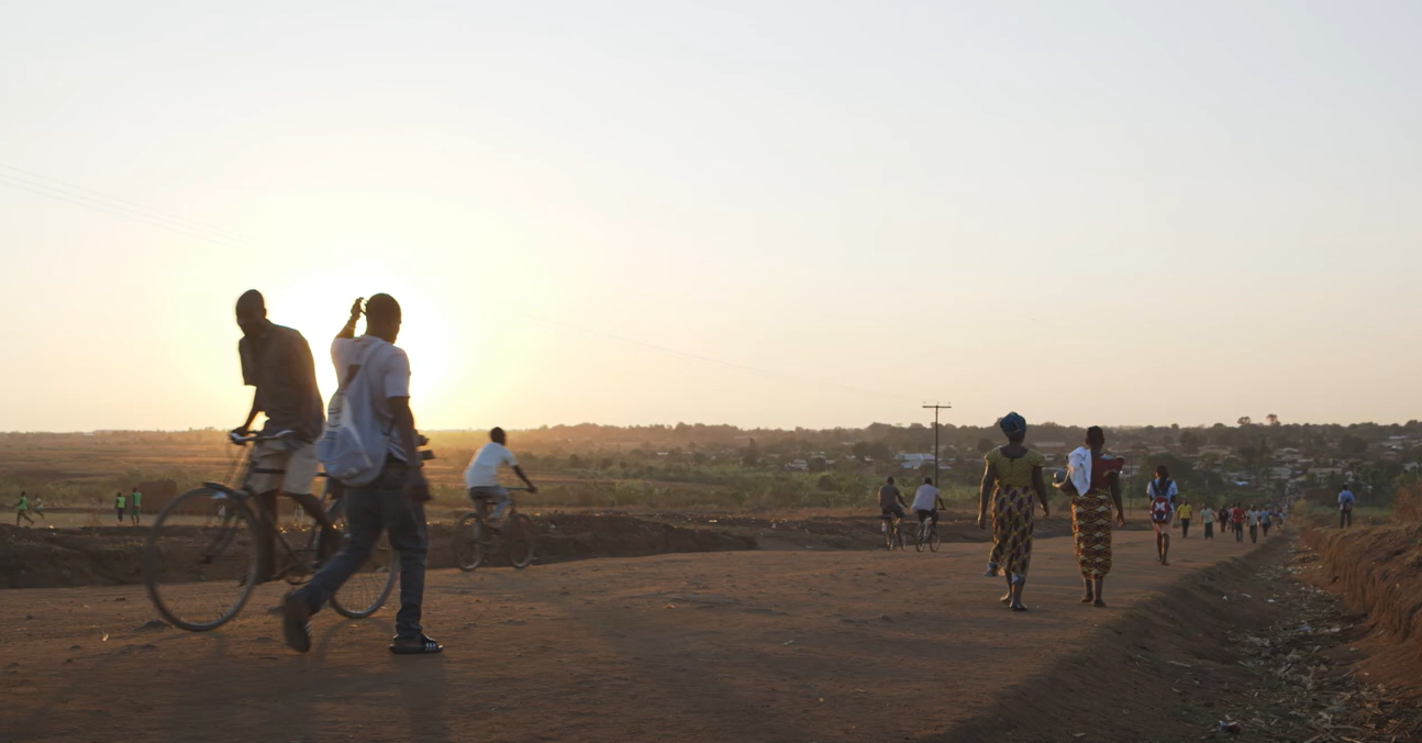 Menschen gehen und fahren mit dem Fahrrad eine staubige Landstraße entlang bei Sonnenuntergang, im Hintergrund ein ländliches Dorf