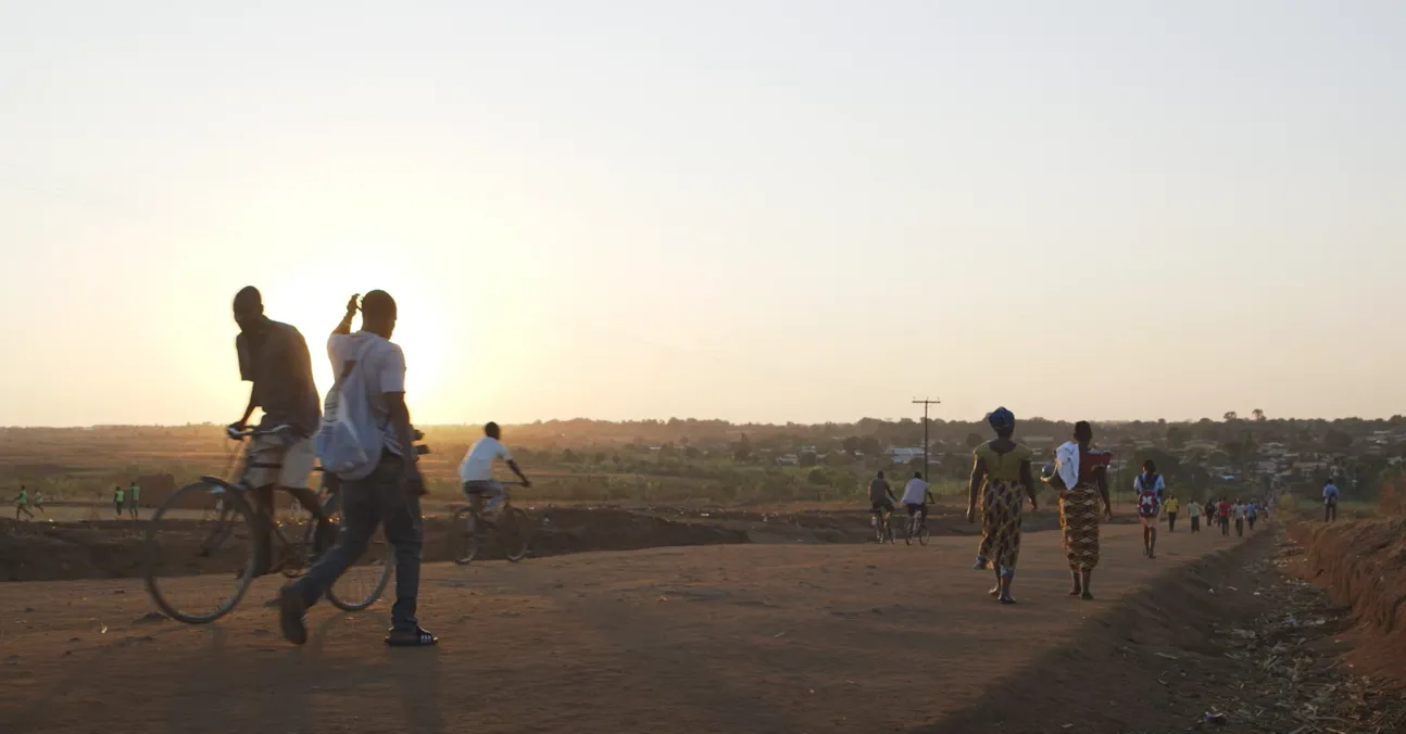 Menschen gehen und fahren mit dem Fahrrad eine staubige Landstraße entlang bei Sonnenuntergang, im Hintergrund ein ländliches Dorf