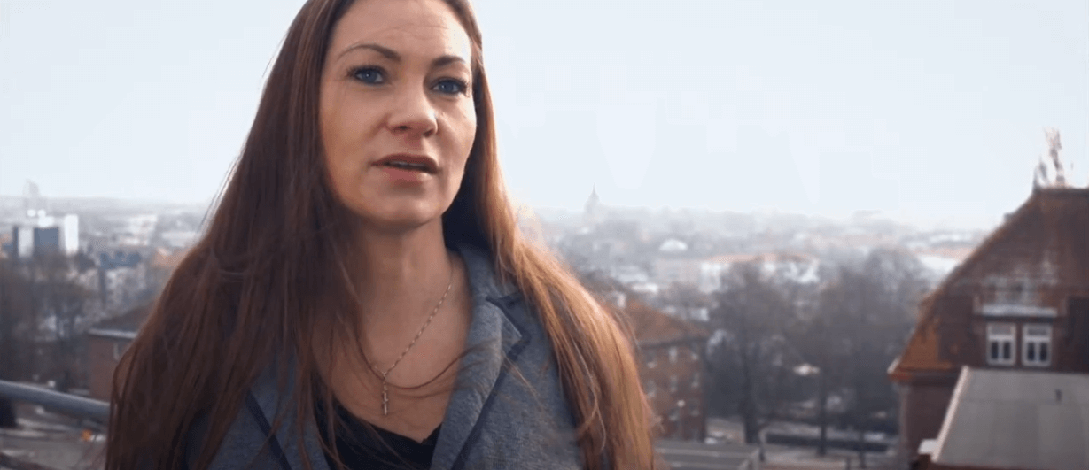 Kvinna som talar på en takterrass med stadsutsikt, iförd grå kavaj och med långt brunt hår.