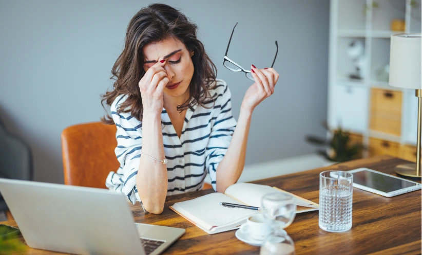 Eine erschöpfte Frau sitzt am Schreibtisch. Sie hat die Augen geschlossen, hält sich den Kopf und ist von Laptop, Tablet und Notizen umgeben.