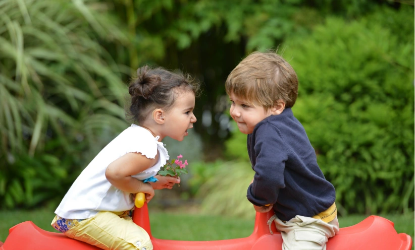 Zwei Kleinkinder sitzen auf einer roten Wippe im Grünen. Das Mädchen mit Blumen spricht, der Bub hört zu.