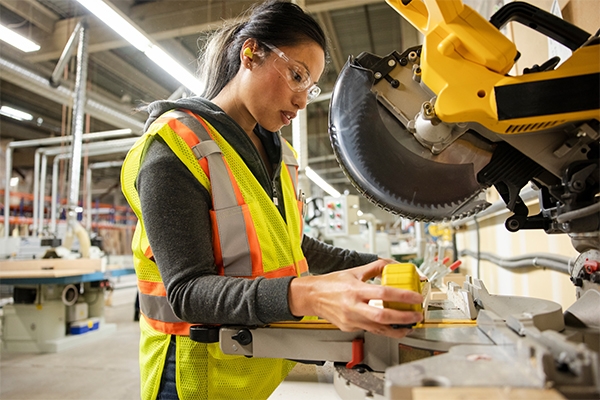 A woman measures and cuts wood while using earplugs for hearing protection.