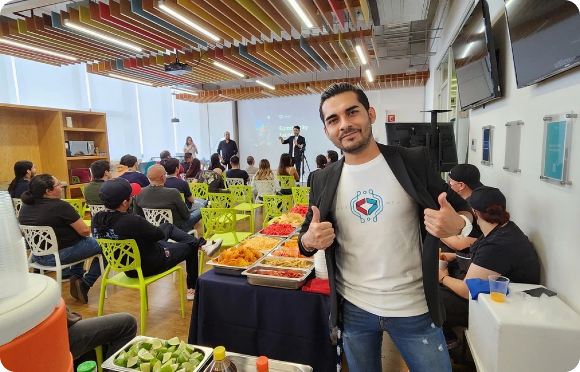 A person in a T-shirt with a hexagon logo is giving a thumbs-up in front of a buffet table at an indoor event where a presentation is taking place in the background.