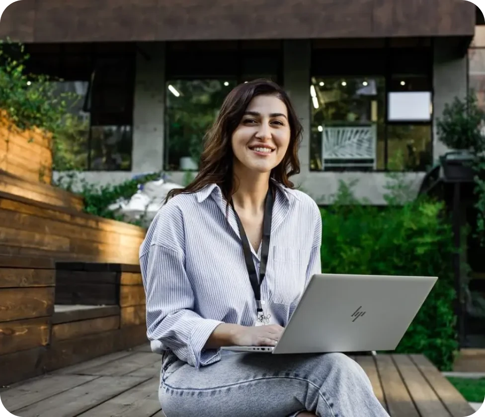A woman sitting on a wooden bench, smiling and looking at her laptop. She is wearing a striped shirt and has a lanyard around her neck, suggesting she might be at a conference or event. The background features a modern building with large windows and greenery.
