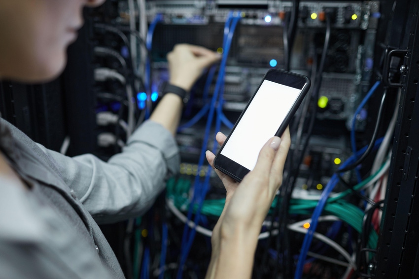 Close up of data center network technician holding smartphone with blank screen while connecting cables in outsourced server cabinet.