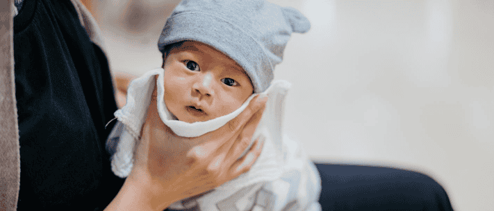 Infant with Gray Hat Held by Adult Against Plain White Wall, with Slightly Surprised Expression