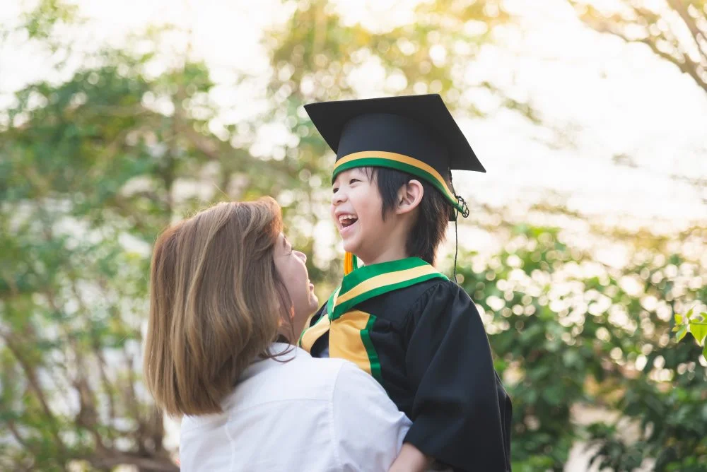 Mom Holding Child In Graduation Cap And Gown, Smiling Against A Background Of Green Trees