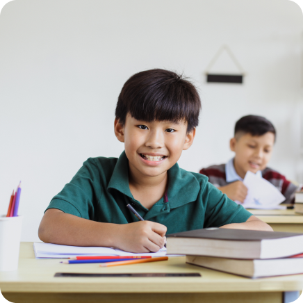 Smiling Preschool Boy Writing On Paper At A Desk Stacked With Books