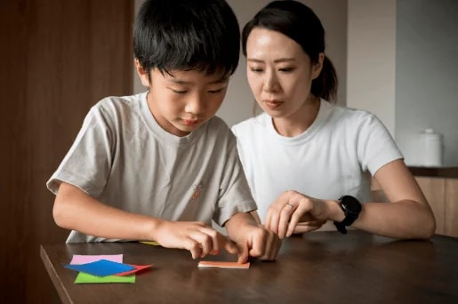 Young Boy And Mother Folding Paper At Dark Wooden Table With Colored Paper Pieces Nearby