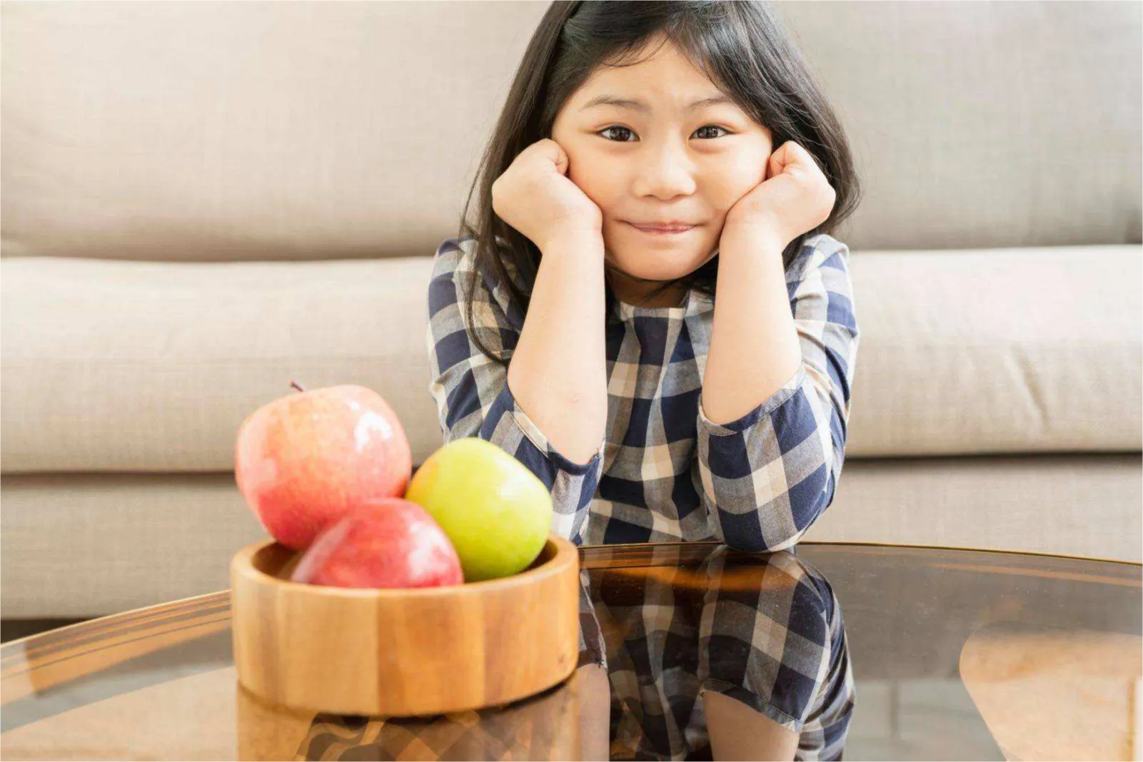 A young girl sits at a glass coffee table with a wooden bowl of red and green apples, hands on her cheeks.