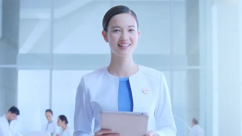 Smiling Woman In White Coat Holding Tablet In Bright Office, With Colleagues In Background
