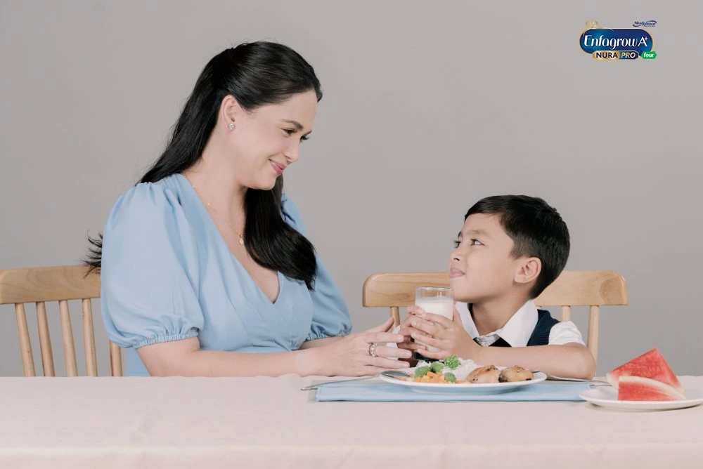 Woman In Light-Blue Dress With Jewelry Smiles At Young Boy Holding Glass Of Milk At A Table With Plates Of Food And Watermelon