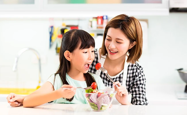 Mother holding a fork to feed daughter salad on the dining table