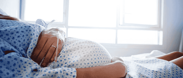 Pregnant Woman in Hospital Bed with IV, Wearing Gown with Blue Flowers, Illuminated by Natural Light from Window