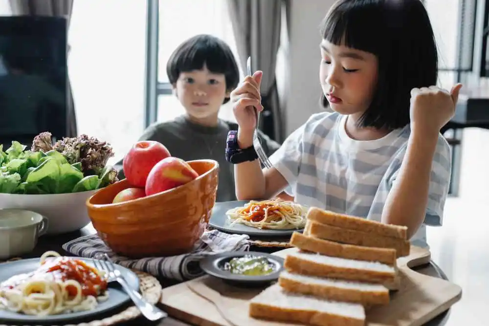 Child Eating Spaghetti With Her Sibling In The Background