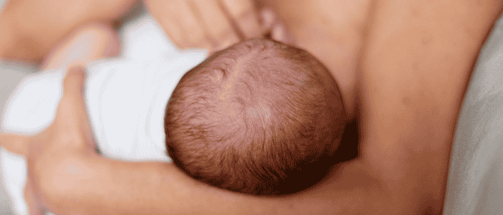 Close-Up of Baby's Head with Brown Hair, Swaddled in White Cloth, Breastfeeding and Held by a Person with Tan Skin