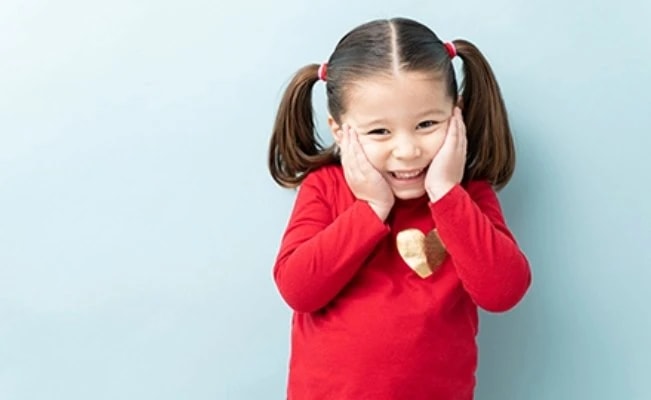 Young girl clasping her cheeks with both hands against plain light blue backdrop