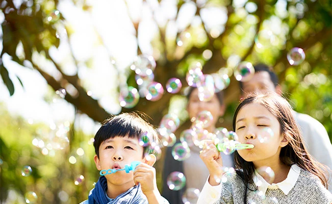 Young children male and female blowing bubbles outdoors with parents in background 