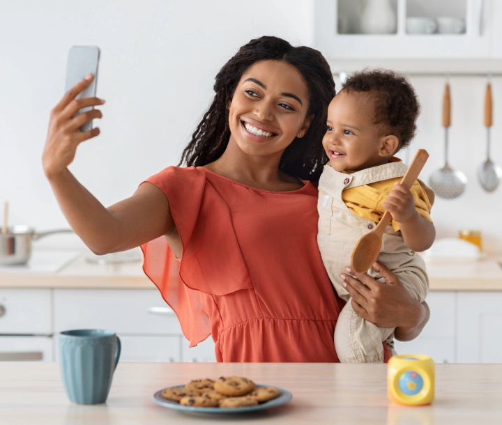 Madre sonriente sosteniendo a su bebé mientras se toman una selfie en la cocina, representando hábitos de alimentación saludable.