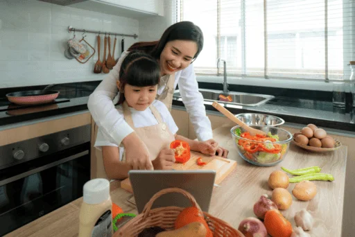 Mother And Daughter Cooking Together In Kitchen, With Tablet And Fresh Produce On Counter