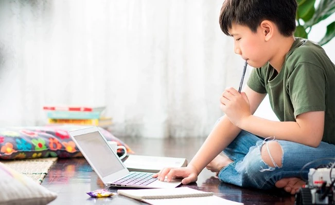 Young boy sitting on floor with laptop and holding a pen, suggesting studying at home