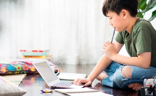 Young boy sitting on floor with laptop and holding a pen, suggesting studying at home