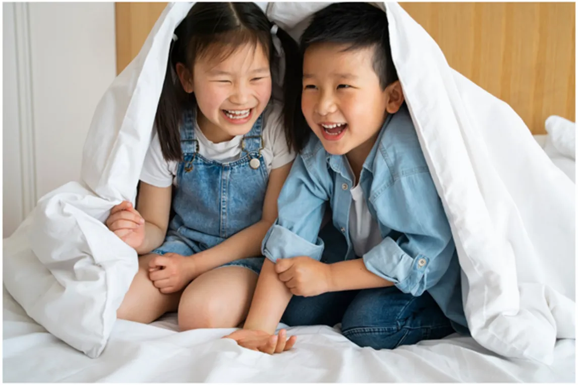 Two-young-children-laughing-together-under-white-blanket-fort.webp