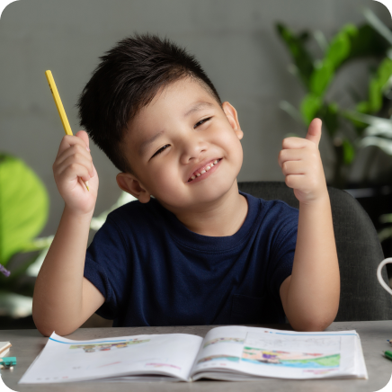 Young boy holding a pen on one hand and another with one thumb up with activity book on desk