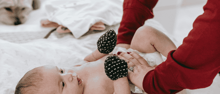 Smiling Infant Lying On Bed While An Adult Gently Changes The Baby’s Diaper, Wearing Polka Dot Socks